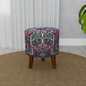 Può includere: Un pouf rotondo e colorato con un motivo geometrico in tonalità di rosa, blu, verde e grigio. Il pouf ha quattro gambe in legno.