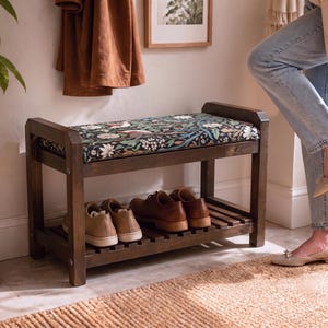 Puede incluir: Un banco zapatero de madera marrón oscuro con un cojín rectangular de asiento con un patrón floral oscuro en tonos verdes, blancos y rosas. El banco incluye un estante inferior de listones para guardar dos pares de zapatos.