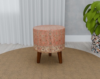 May include: A round, pink and cream patterned ottoman with wooden legs. The ottoman is sitting on a beige rug.