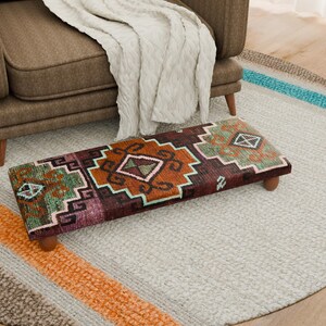 Puede incluir: Un reposapiés rectangular con un diseño estampado en tonos marrón, naranja, verde y morado. El reposapiés tiene patas de madera y está colocado sobre una alfombra gris claro. Un sillón marrón y una manta color crema están en el fondo.
