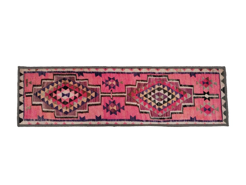 May include: A rectangular, pink runner rug with geometric patterns. The rug features diamond and triangle shapes in shades of pink, purple, black, and beige. A gray border frames the edges.