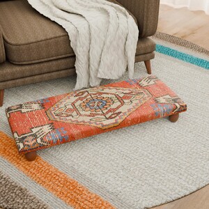 May include: A rectangular footstool with a patterned rug design in red, orange, blue, and cream. The footstool has wooden legs and sits on a light grey rug. The rug is near a brown armchair and a white blanket. The footstool's dimensions are approximately 60 cm long.