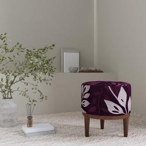 May include: A round, upholstered ottoman with a dark purple background and white leaf pattern. The ottoman has a wooden base with four legs. A vase with green branches and a small diffuser are in the background.
