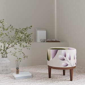 Peut inclure: Un pouf rond décoratif avec un motif botanique. Le tissu présente des motifs de feuilles violettes et blanches sur un fond beige. Il a une base et des pieds en bois foncé. Un vase avec des branches vertes et des livres sont à proximité.