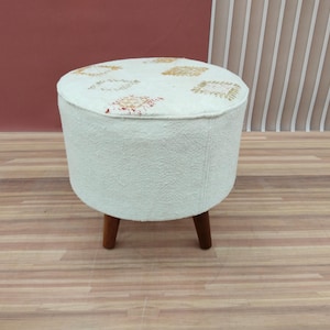 May include: A round, off-white ottoman with a patterned top and three wooden legs. The top features a repeating design in shades of gold and red. The ottoman is a decorative furniture piece.