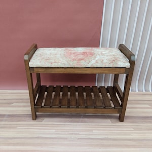 Puede incluir: Un banco de madera con un asiento acolchado y un estante inferior. El cojín está cubierto con una tela estampada en rojo, blanco y beige.