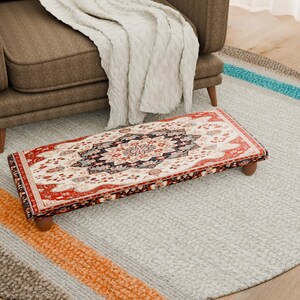 Puede incluir: Un reposapiés rectangular con un diseño de alfombra detallado en rojo, crema y azul marino. El reposapiés tiene patas cortas y redondeadas de madera. El patrón de la alfombra presenta un medallón central y motivos florales.