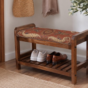 Puede incluir: Un banco zapatero de madera con marco rectangular y estante inferior de listones. El asiento superior cuenta con un cojín de tela color óxido con un patrón repetitivo de criaturas estilizadas en beige y marrón oscuro y motivos florales.