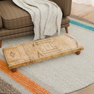 Puede incluir: Un reposapiés rectangular de madera envejecida con un patrón geométrico se encuentra sobre una alfombra gris claro. El reposapiés tiene cuatro patas pequeñas y redondeadas de madera. Un sillón tapizado y una manta color crema están en el fondo.