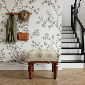 a wooden ottoman with a beige cushion, positioned in front of a wall with a black and white floral wallpaper. A coat rack is mounted on the wall above the ottoman, and a hat is hanging on the wall. A staircase is visible in the background.
