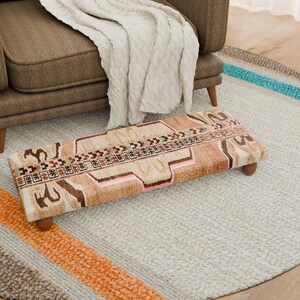 Puede incluir: Un otomán rectangular con un diseño estampado multicolor, con tonos marrones, rosas y beige. El otomán descansa sobre cuatro patas cortas de madera. Un sillón marrón y una manta color crema están en el fondo, junto con una alfombra redonda.