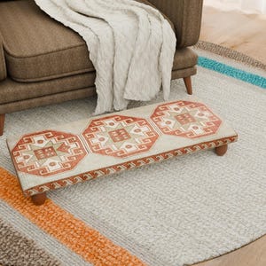 Puede incluir: Un reposapiés rectangular con un patrón geométrico beige y rojo se encuentra sobre una alfombra gris claro. El reposapiés tiene patas de madera y una parte superior estampada. Un sillón marrón y una manta blanca están en el fondo.