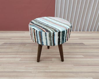 May include: A round, striped ottoman with a white, brown, blue, and pink pattern. The ottoman has four wooden legs.