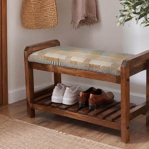 May include: A wooden entryway bench with a rectangular seat cushion featuring a multi-colored plaid pattern in shades of tan, blue, and cream. The bench has a lower slatted shelf for storage and a dark brown wood finish with visible grain.