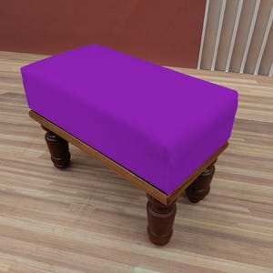 May include: A rectangular purple ottoman with a wooden frame and four dark brown, turned legs. The ottoman is a solid color and sits on a light brown wooden floor.