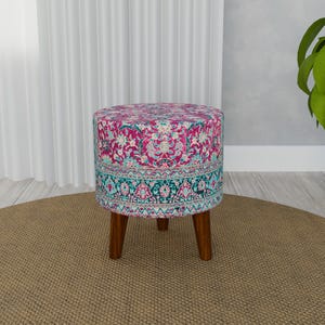 May include: A round, pink and blue patterned ottoman with wooden legs. The ottoman is sitting on a brown rug.