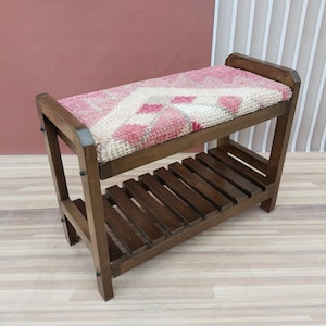 May include: Wooden bench with a woven seat cushion featuring a pink and cream geometric pattern. The bench has a lower shelf made of wooden slats and is constructed with dark brown wood. The design is simple and functional.
