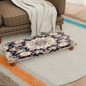 Puede incluir: Un reposapiés rectangular con una tela estampada con fondo azul marino y diseños florales en crema, rosa y azul. El taburete tiene patas de madera y está colocado sobre una alfombra gris claro cerca de un sillón marrón. Mide unos 76 cm de largo.