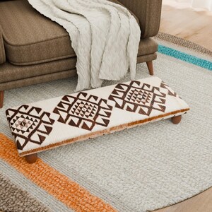 Puede incluir: Un reposapiés rectangular con fondo crema y un patrón geométrico marrón. El reposapiés tiene patas de madera y está sobre una alfombra gris claro cerca de un sillón marrón y una manta crema. La alfombra tiene un diámetro de aproximadamente 150 cm.