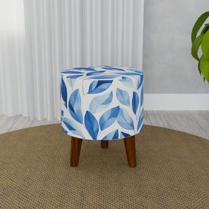 Può includere: Un pouf rotondo blu e bianco con motivo floreale e gambe in legno. Il pouf è su un tappeto marrone.