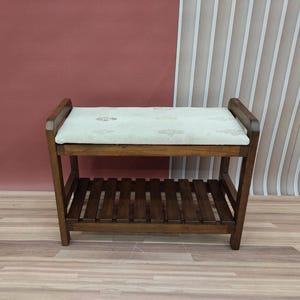 Può includere: Una panca in legno con una seduta imbottita e uno scaffale sottostante. La panca è marrone e il cuscino è bianco con un motivo floreale.