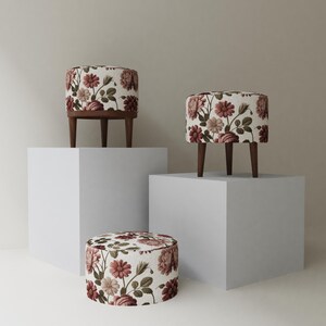 May include: Three floral patterned ottomans with dark wooden legs. The ottomans have a cream-colored background with burgundy, pink, and green floral designs. One ottoman is on the floor, and two are on white pedestals.