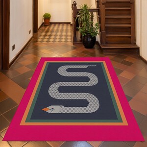 May include: A rectangular rug featuring a geometric snake design. The rug has a hot pink border, followed by orange and green stripes, and a navy blue centre. The snake is white with a grey geometric pattern and an orange head.