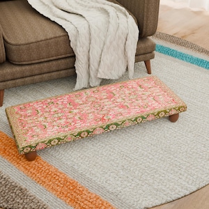 Puede incluir: Un reposapiés rectangular rosa con un estampado floral y ribete verde, apoyado sobre cuatro patas de madera. El reposapiés está colocado sobre una alfombra gris claro con rayas coloridas, cerca de un sillón marrón y una manta blanca.