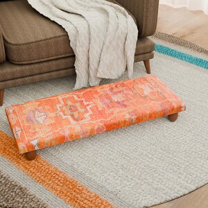 Puede incluir: Un reposapiés rectangular con un estampado naranja y rosa, con patas de madera, colocado sobre una alfombra gris claro. Un sillón marrón y una manta crema están en el fondo. La alfombra tiene rayas de colores.