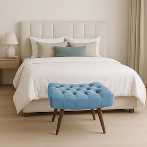Puede incluir: Un otomán azul claro con mechones y patas de madera se encuentra frente a una cama con cabecero crema y ropa de cama blanca. La habitación tiene una paleta de colores neutros, con una mesita de noche y una lámpara a la izquierda.
