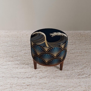 May include: An upholstered ottoman with a dark blue wave pattern and gold accents. The base of the ottoman features a fan-like design in teal and gold. It has three wooden legs and is placed on a light-colored rug.