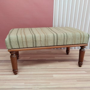 Puede incluir: Un otomano de tela a rayas con un marco de madera y cuatro patas torneadas. El otomano está sobre un suelo de madera.