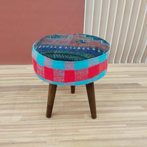 Puede incluir: Un otomano redondo y colorido con estampado y tres patas de madera. El otomano está cubierto con un mosaico de tela roja, azul y negra.