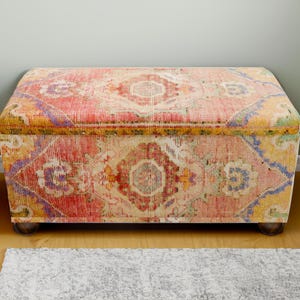 Può includere: Un pouf rettangolare con un rivestimento in tessuto fantasia colorato. Il tessuto presenta un mix di tonalità rosse, rosa, gialle e viola in un design geometrico. Il pouf poggia su quattro piccoli piedi in legno scuro.