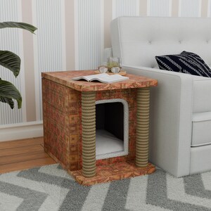 Può includere: Una cuccia per gatti a forma di piccolo mobiletto in legno decorato con un bordo in corda. Il mobiletto ha un design a motivi con colori rosso, arancione e marrone. La cuccia per gatti è posizionata su un tappeto bianco e grigio davanti a un divano bianco.