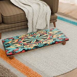 Puede incluir: Un reposapiés rectangular con un diseño colorido e intrincado con patrones turquesa, naranja y negro. El taburete tiene cuatro patas cortas de madera y está colocado sobre una alfombra gris claro. Un sillón marrón y una manta blanca están en el fondo.