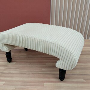 Puede incluir: Otomana de pana color crema con un diseño curvo y cuatro patas torneadas negras. La tela texturizada y la forma curva le dan un aspecto clásico y elegante. Este reposapiés es una adición elegante a cualquier espacio habitable.