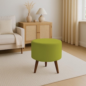 Pode incluir: Um pufe redondo verde limão com três pernas de madeira castanha escura. O assento estofado é verde brilhante liso. O pufe está colocado sobre um tapete creme, sugerindo um design de interiores moderno. Tem cerca de 40 cm de altura.