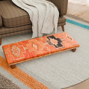Puede incluir: Un reposapiés rectangular con un patrón naranja y negro, sostenido por patas de madera. Descansa sobre una alfombra gris claro, colocado frente a un sillón marrón. Una manta crema está sobre la silla.