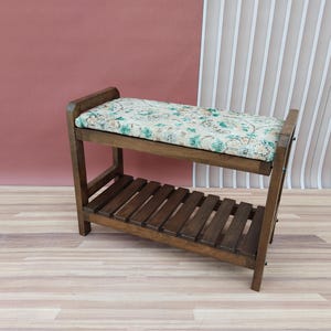 May include: A wooden bench with a cushioned seat and a lower shelf. The bench is brown and the cushion has a floral pattern.