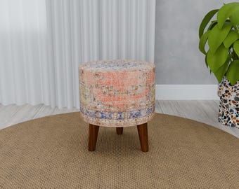 Pode incluir: Um pufe redondo estampado com um design rosa, azul e branco. O pufe tem quatro pernas de madeira e está sobre um tapete castanho.