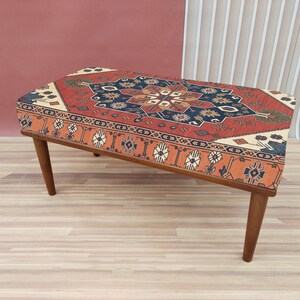 Può includere: Un pouf in legno con un top in tessuto a motivi rossi, arancioni, blu e bianchi. Il pouf ha un design tradizionale e sarebbe un'ottima aggiunta a qualsiasi casa.
