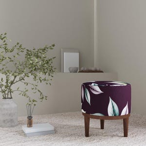 May include: A circular ottoman with a dark purple fabric featuring a white and green leaf design. It has a wooden base and legs. The room also includes a vase with foliage and a decorative shelf with objects.