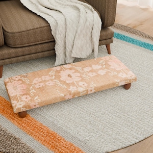 May include: A rectangular footstool with a floral pattern in shades of pink and beige, resting on four wooden legs. The footstool is placed on a round rug with a light gray background and colorful stripes.