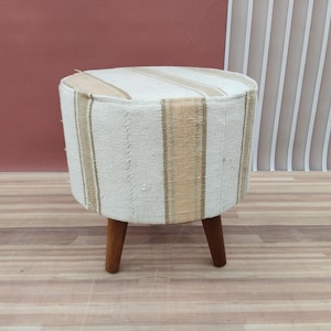 Op de afbeelding: Een ronde, gestoffeerde poef met een crèmekleurig en beige gestreept patroon. De poef heeft drie donkere houten poten. De neutrale kleuren en het eenvoudige ontwerp maken het een veelzijdig meubelstuk. Hoogte: ongeveer 40 cm.