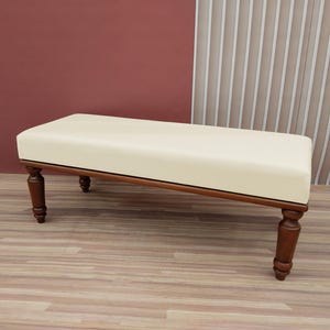 May include: A white upholstered bench with wooden legs. The bench has a simple design and is perfect for adding a touch of elegance to any room.