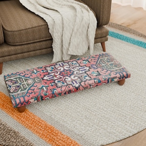 May include: A rectangular footstool with a pink, blue, and white patterned design. The footstool has wooden legs and is placed on a light gray rug. A brown armchair and a cream-colored blanket are in the background.