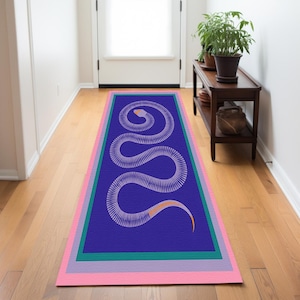 Puede incluir: Una alfombra rectangular con un diseño de serpiente blanca estilizada sobre un fondo azul. La alfombra está bordeada por rayas verdes, moradas y rosas. La cabeza de la serpiente está en la parte superior y su cola se curva en la parte inferior.