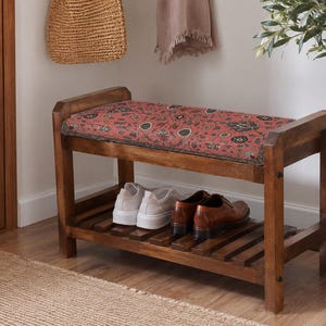 May include: A wooden shoe bench with a rectangular frame and a slatted lower shelf. The top features a cushioned seat upholstered in a red fabric with a dark, intricate floral and paisley pattern. Two pairs of shoes are placed on the bottom shelf.