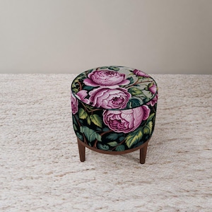 May include: A round ottoman with a floral pattern featuring large pink roses on a black background. The ottoman has a dark wooden base and three short legs. The fabric design includes green leaves and stems.